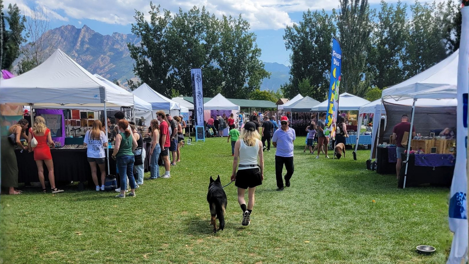 A German Shepherd heeling calmly on a leash through a busy Utah outdoor market with tents and mountains in the background